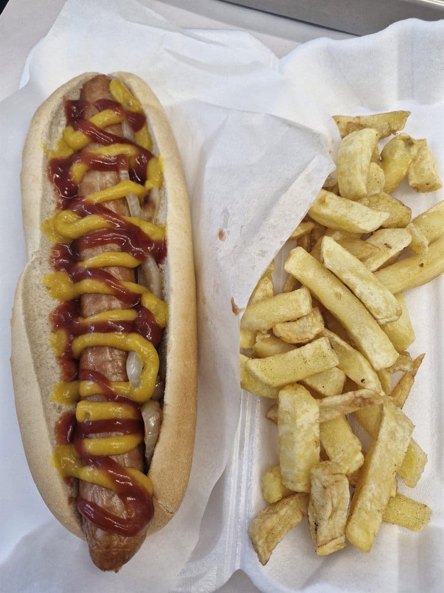 A Bigfoot sausage in a long bun topped with onions, ketchup and mustard, served with a portion of chips
