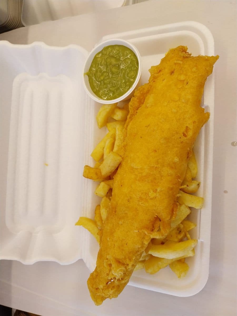 Battered fish on a bed of chips in a takeaway box, with a pot of mushy peas alongside